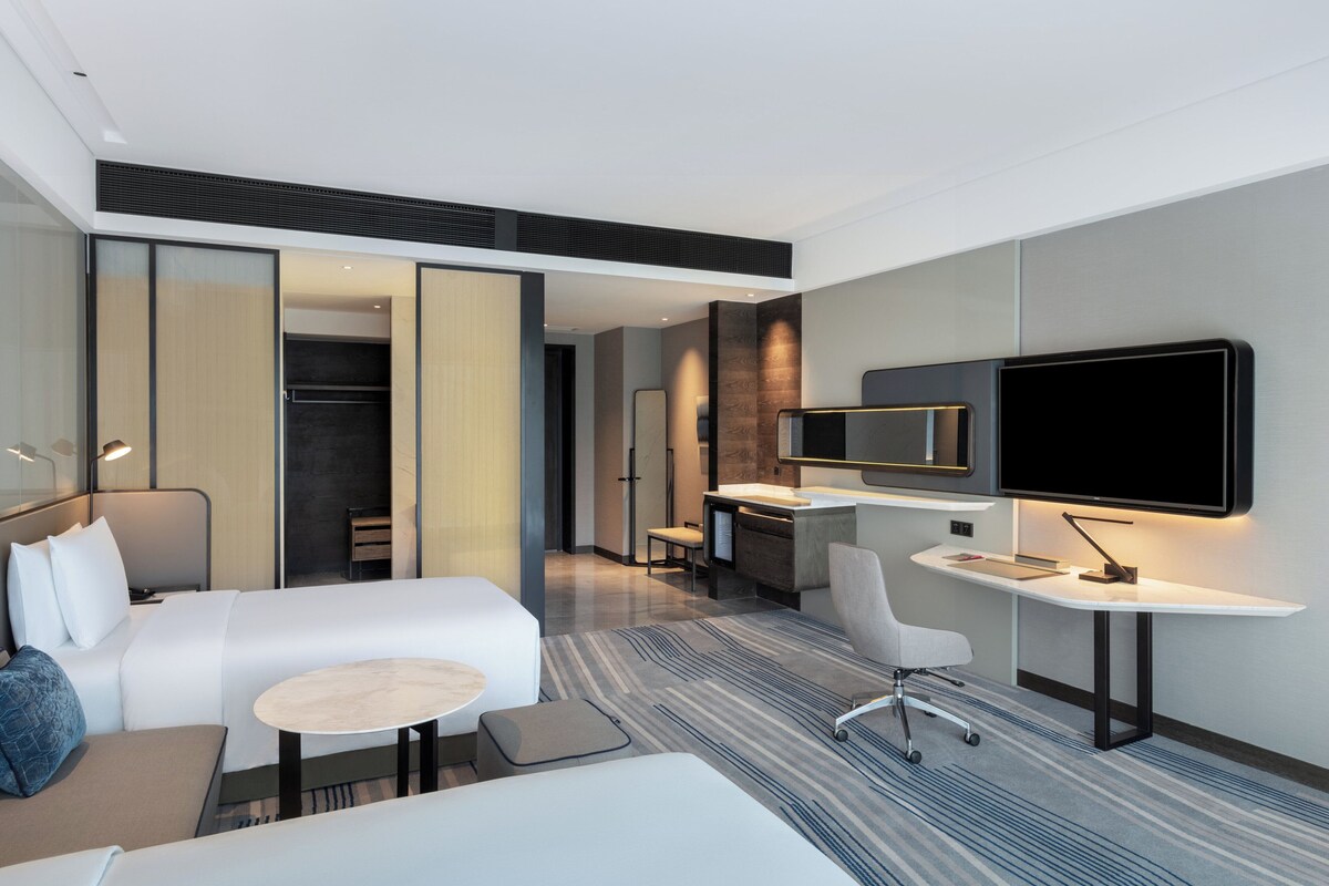 A modern hotel room with a large bed, a desk, a chair, a TV, and a walk-in closet.