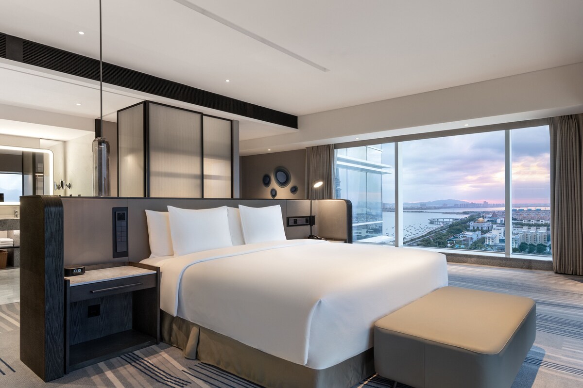 A modern hotel room with a large bed, a desk, and a view of the cityscape.