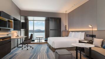 Ein modernes Hotelzimmer mit einem großen Bett, einem Schreibtisch, einem Sessel, einer Couch, einem Fernseher und Blick ins Freie.