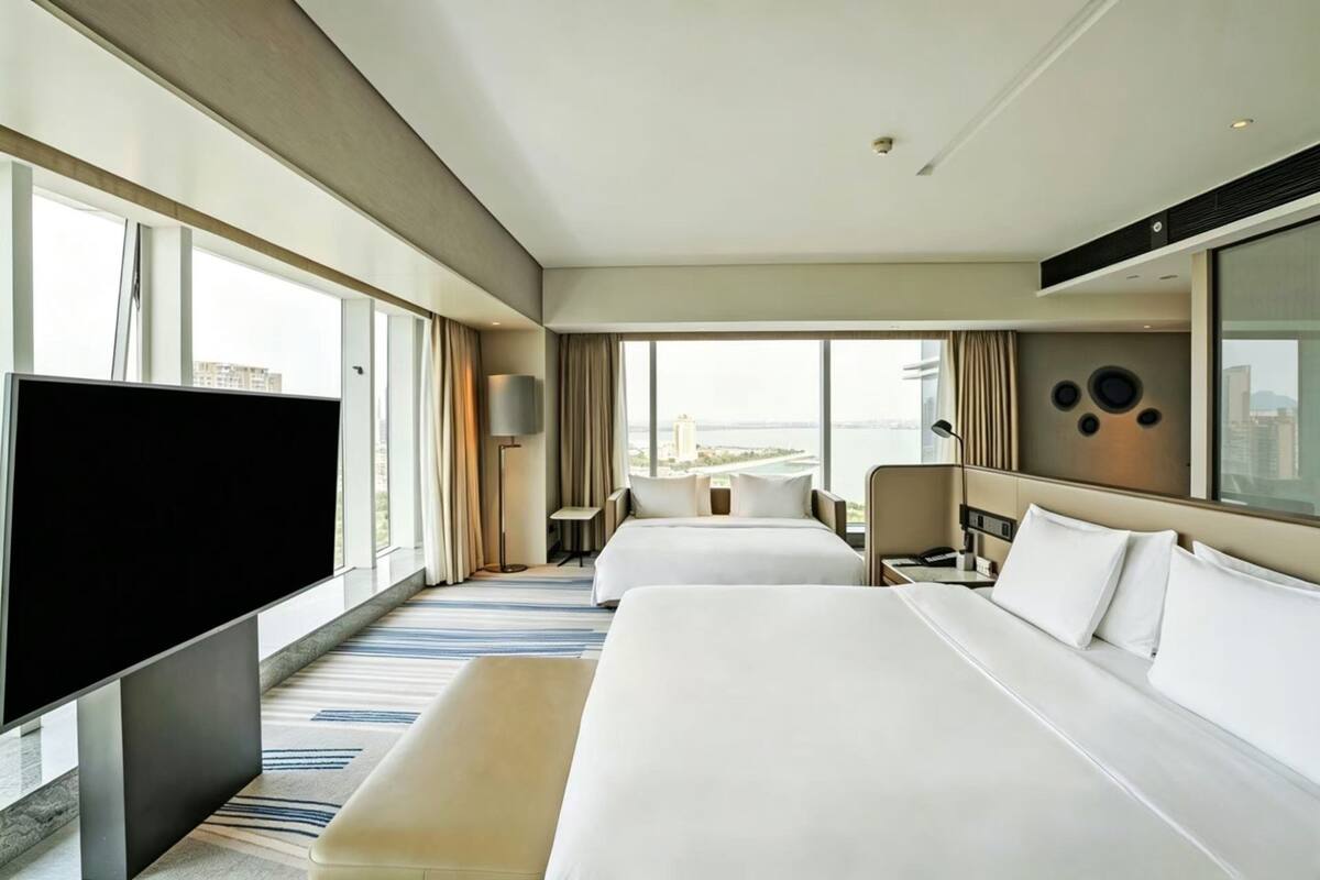 A hotel room with two beds, a flat-screen TV, a large window with a city view, and a modern design.