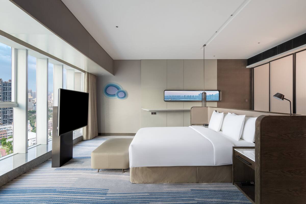A modern hotel room with a large bed, a flat-screen TV, a city view, and a minimalist design.