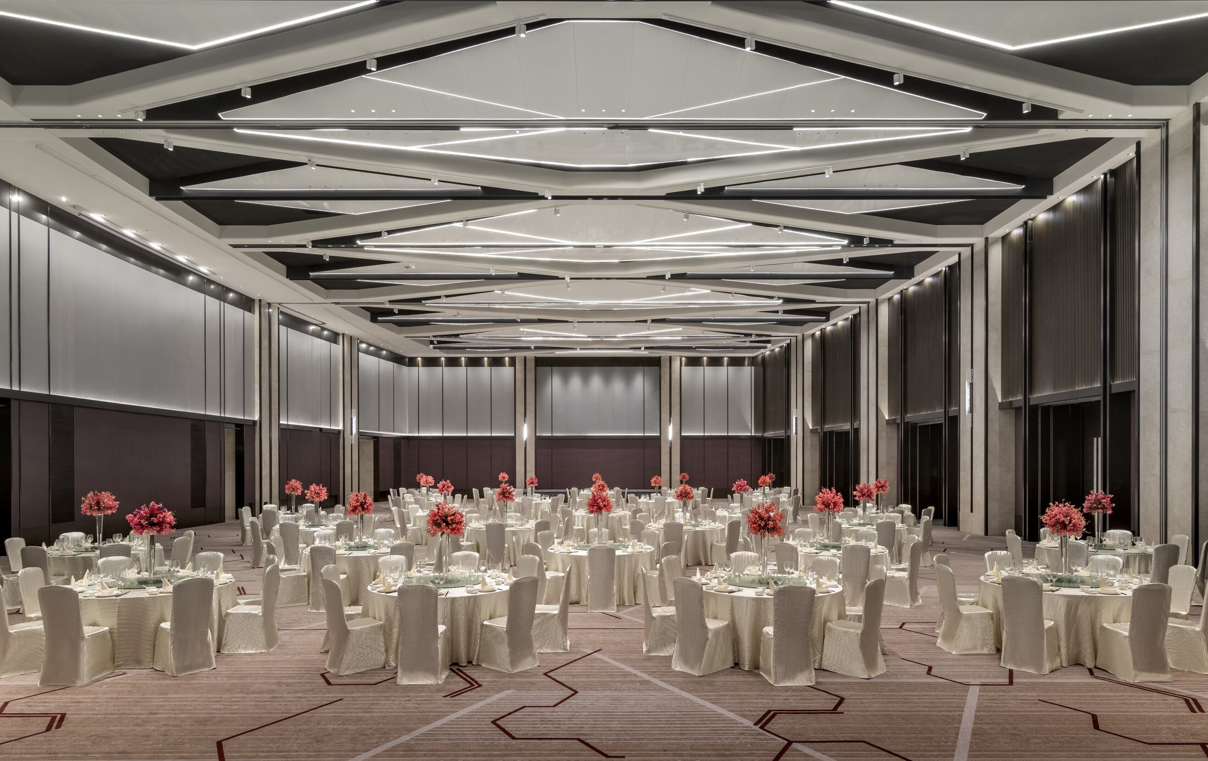 Una amplia sala de conferencias con varias mesas redondas, cada una cubierta con manteles blancos y sillas, y centros de mesa con flores rojas.