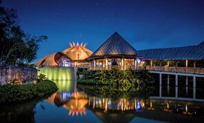 Experience the serene ambiance of our resort, where a unique dome-shaped building and a pyramid-shaped building are connected by a bridge over a tranquil body of water, surrounded by lush greenery.