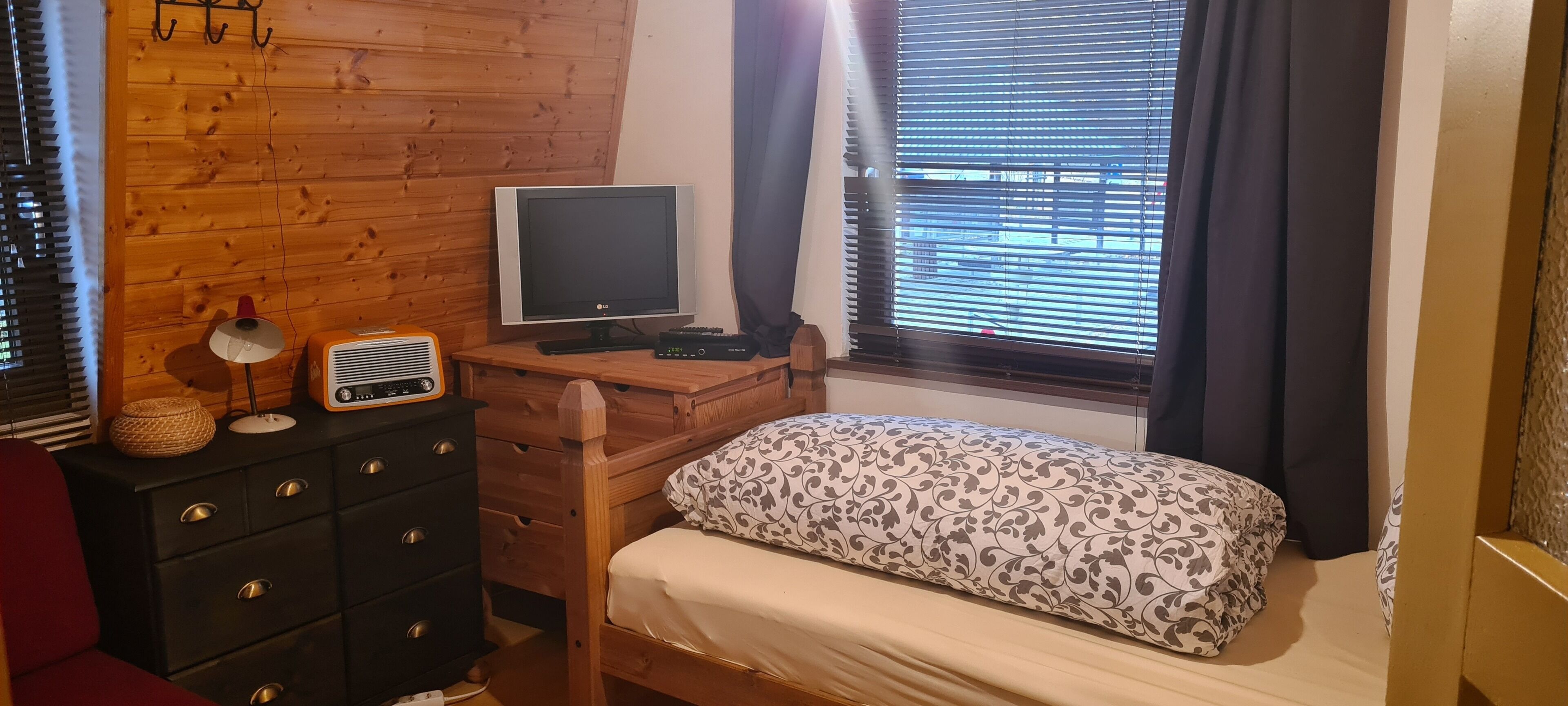 Une chambre avec un bureau en bois, une télévision, une radio, un lit avec une literie à motifs et une fenêtre avec des stores.