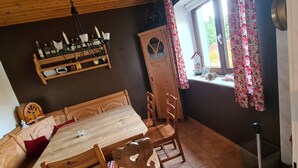 A dining area with a wooden table and chairs, a wooden cabinet with a shelf above, and a window with floral curtains.