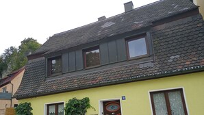A two-story house with a yellow facade, a brown door, and a small garden area with climbing plants.