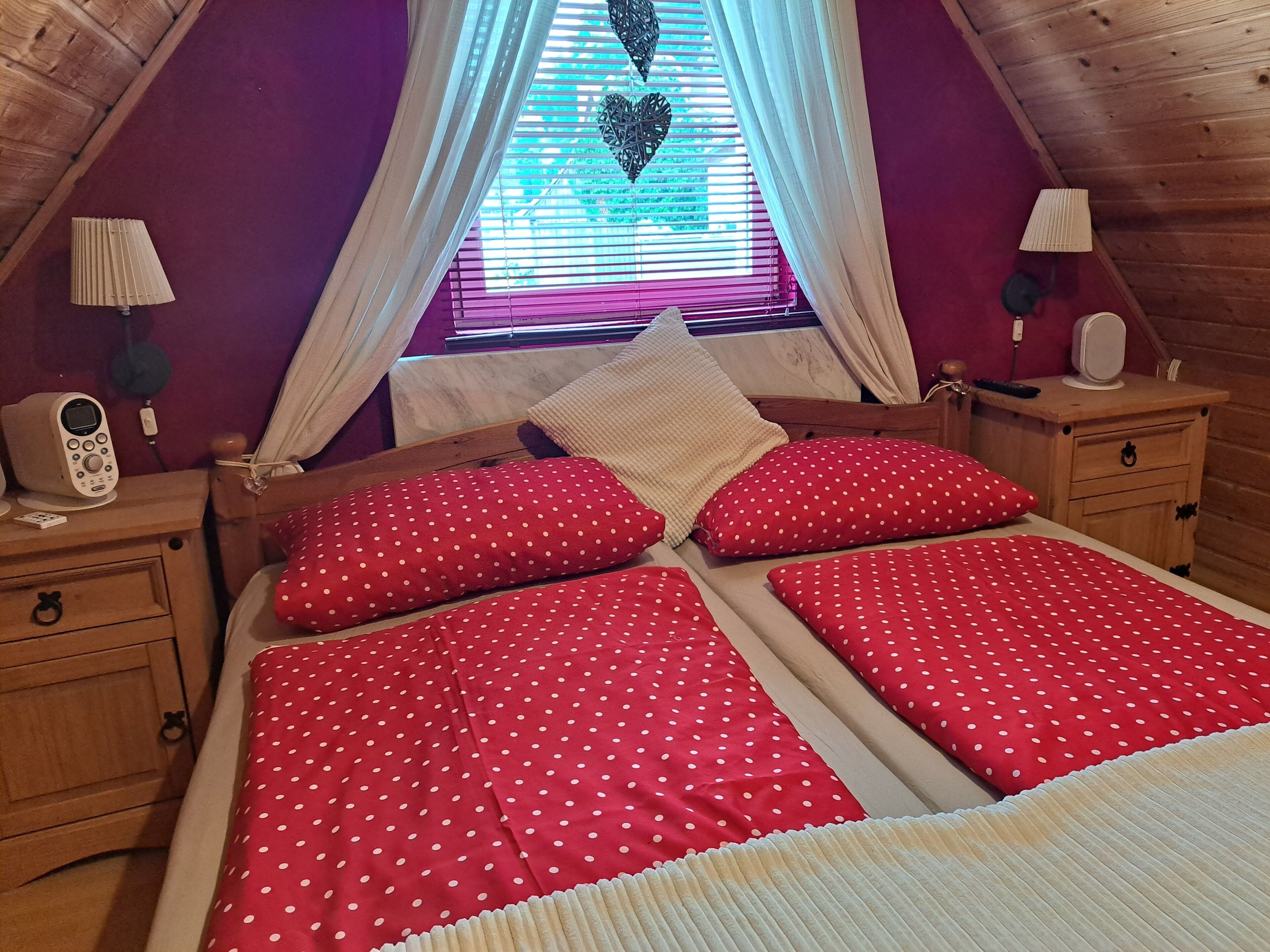 A cozy attic bedroom with a sloped ceiling, a bed with red polka dot bedding, two bedside tables with lamps, and a window with a heart-shaped decoration.