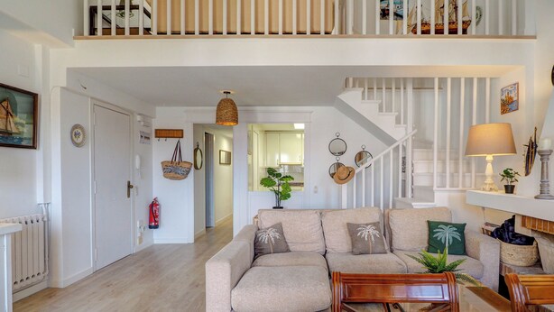 En rummelig opholdsstue med sofa, sofabord og spisebord dækket til fire personer. En trappe fører op til en etage ovenpå.