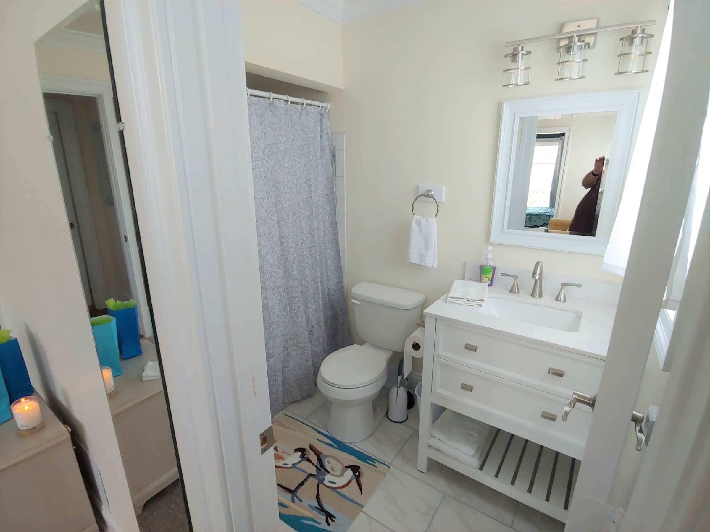 Een badkamer met toilet, wastafel en spiegel. Er is een douchegordijn, een handdoekhouder en een kleine prullenbak.
