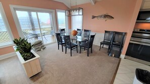 A dining area with a glass table and chairs, a fish decoration on the wall, and a view of the outdoors through large windows. - 6812 Beach Rd, Semi-Oceanfront/Pool (Nags Head)