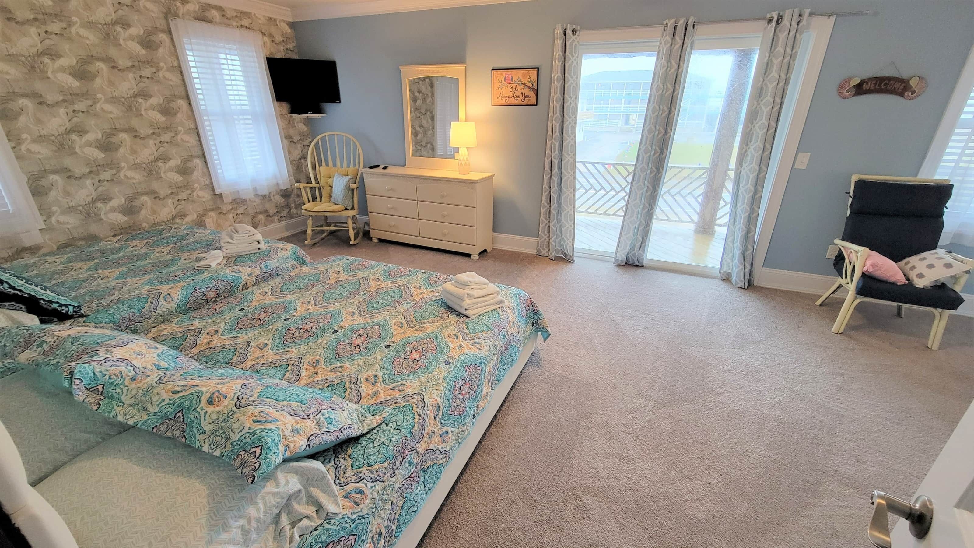 A bedroom with a large bed, a dresser, a chair, a TV, and a balcony with curtains.