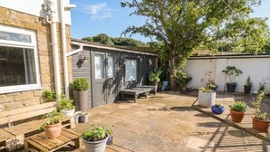 A patio with wooden tables and potted plants, a grey building with a window, and a tree providing shade. - Asta Cottage (Morpeth)