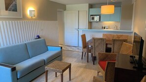 Ein modernes Wohnzimmer mit einer Couch, einem kleinen Tisch und Korbstühlen. Es gibt eine Küchenzeile mit einer Mikrowelle und einer Hängeleuchte.