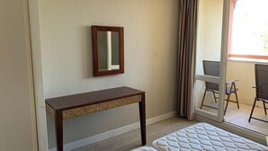 Ein Zimmer mit einem Bett, einem kleinen Holztisch, einem Spiegel und einer Glastür, die zu einem Außenbereich mit Sitzgelegenheiten führt.