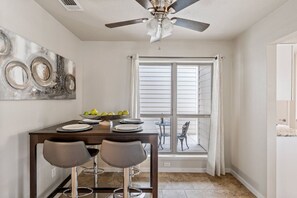 A dining area with a table, two bar stools, a ceiling fan, and a window with blinds.