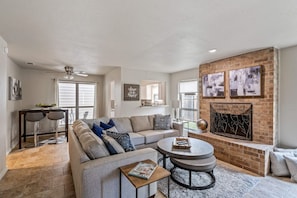 A spacious living room with a grey sofa, a round coffee table, and a fireplace. There are two bar stools and a dining table with chairs. - Single Story Home. Walk to shops and restaurants (Bedford)