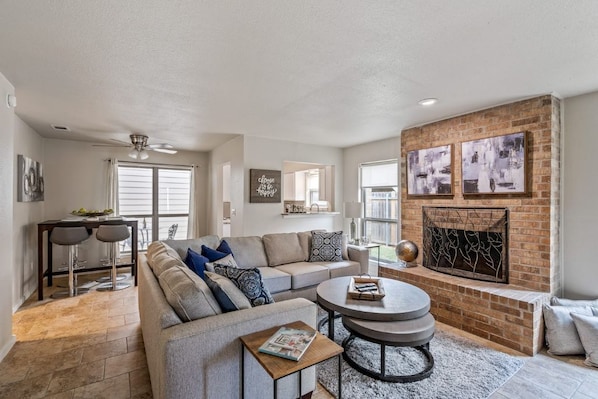 A spacious living room with a grey sofa, a round coffee table, and a fireplace. There are two bar stools and a dining table with chairs.