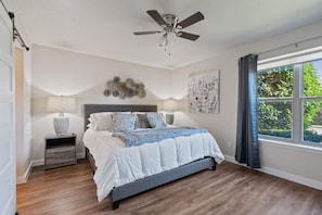 Ein Schlafzimmer mit Bett, Nachttischlampen, Deckenventilator, Fenster mit Vorhängen und einem Wandbild.