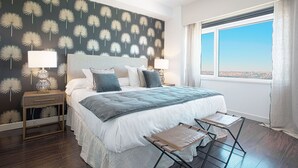 Une chambre moderne avec un grand lit, deux tables de chevet avec des lampes, une vue sur la ville par une grande fenêtre et un mur orné d’un motif de pissenlit.