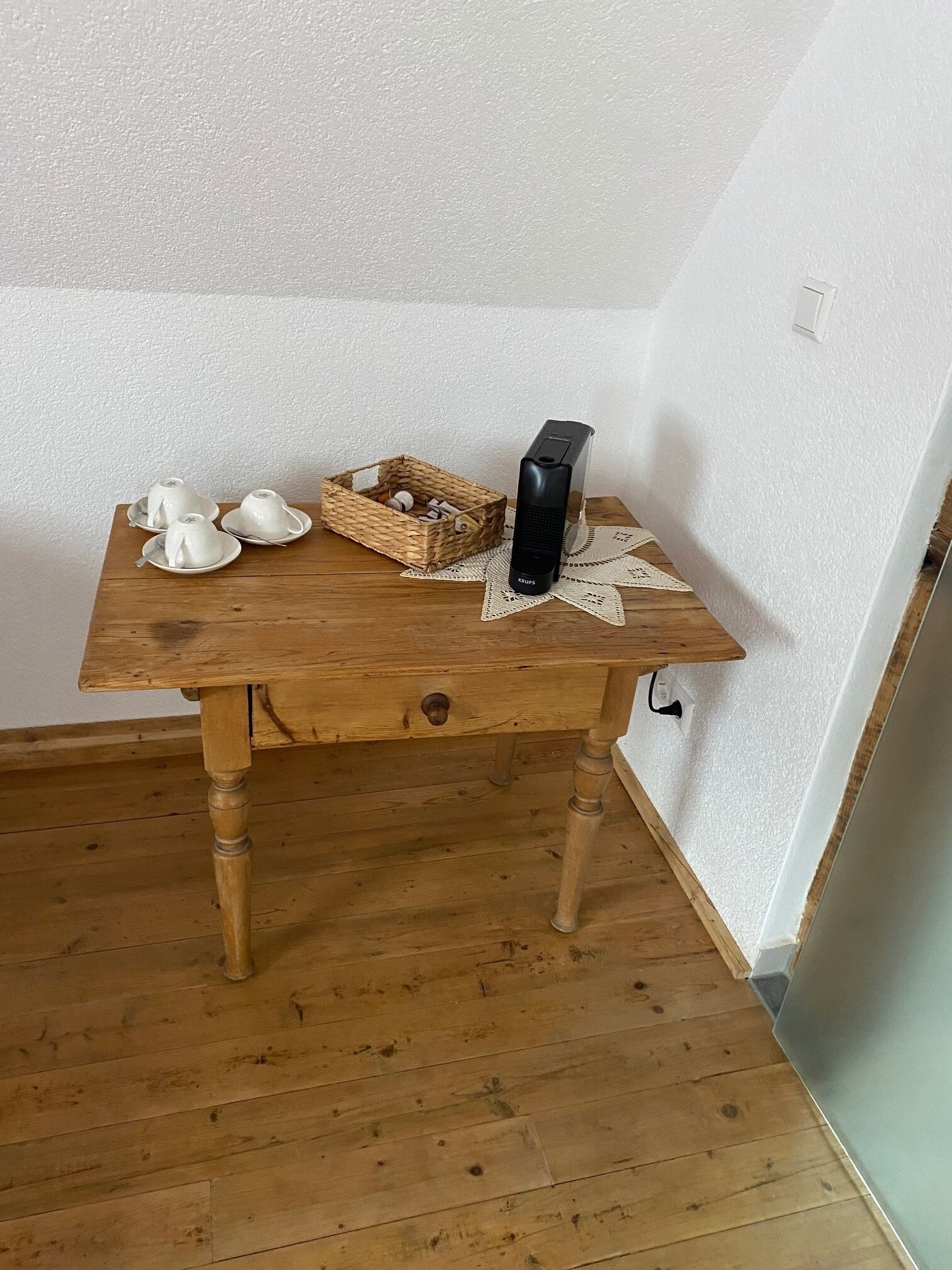 Une table en bois sur laquelle sont posés un panier tressé, trois théières blanches et une bouilloire électrique noire, le tout reposant sur un chemin de table en dentelle.