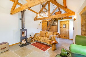 Una sala de estar acogedora con techo de madera, un sofá, un sillón verde, una chimenea y una escalera que lleva a un altillo.