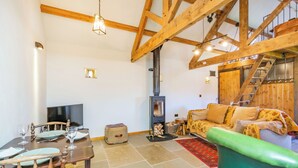 A spacious living area with a high ceiling, wooden beams, a fireplace, a sofa, a dining table set, and a television.