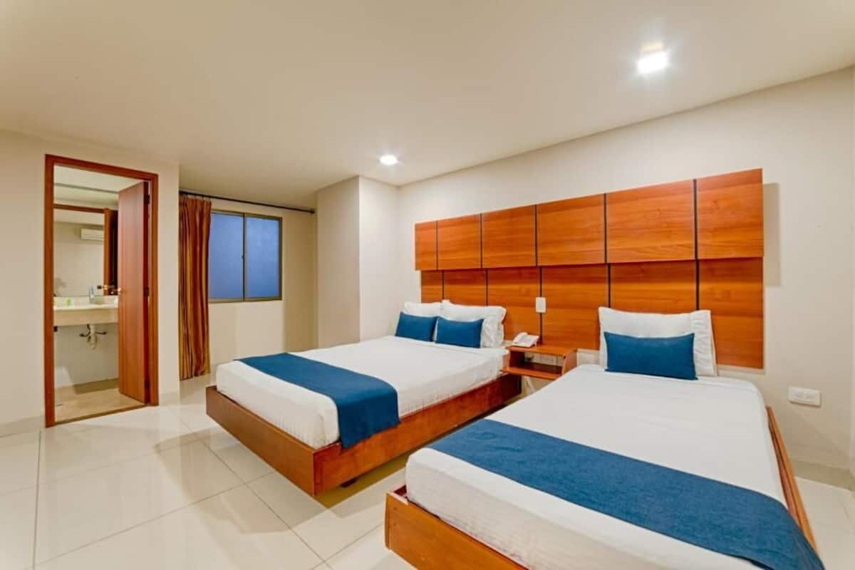 A hotel room with two beds, a wooden headboard, a bedside table with a lamp, and a bathroom visible through an open door.