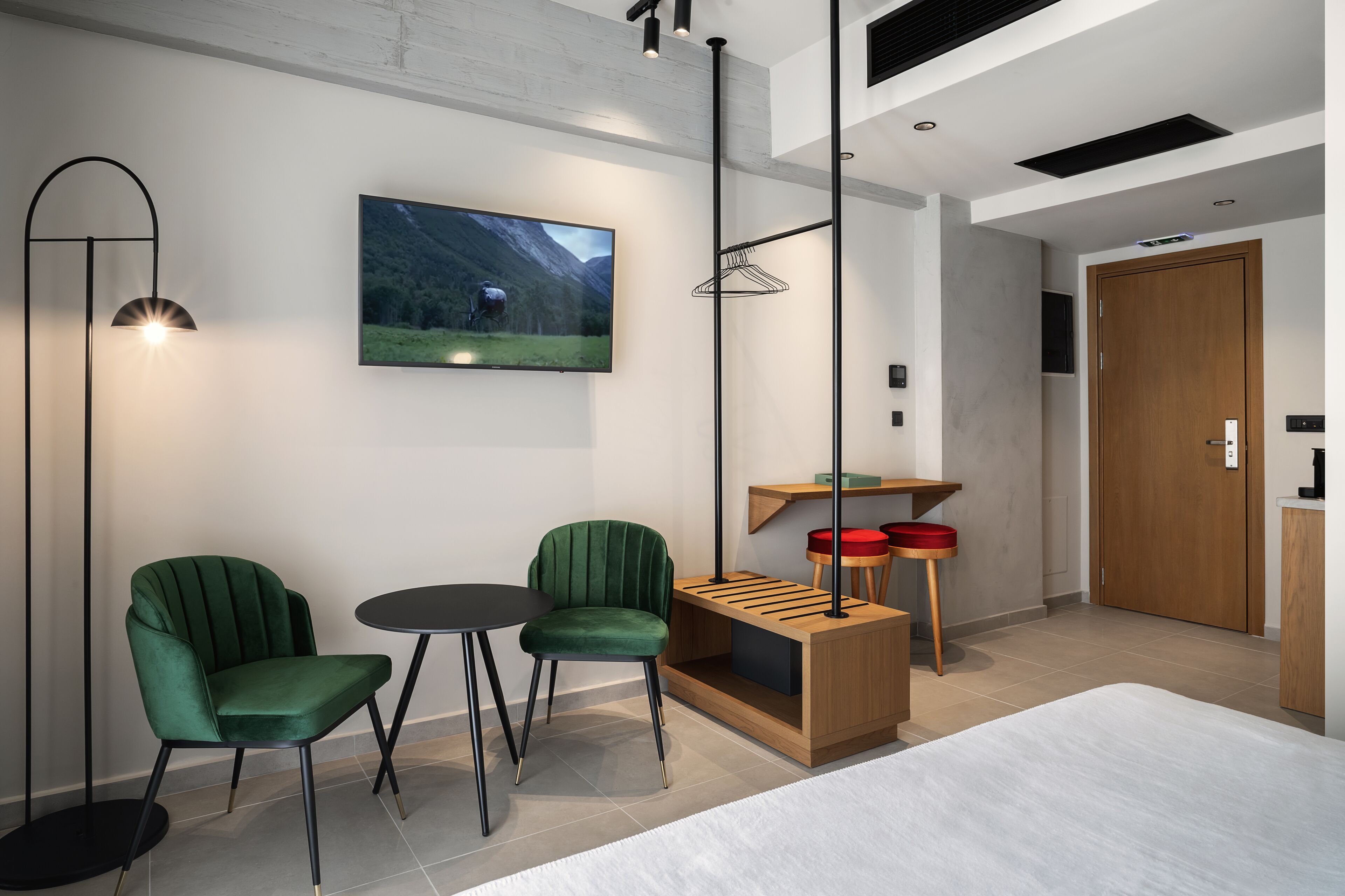 Una camera d'albergo moderna con un letto, due poltrone rivestite di verde, un piccolo tavolino, una TV a parete e una lampada a sospensione.
