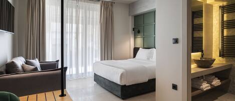 A modern hotel room with a bed, a seating area, and a bathroom with a vanity and towel rack.