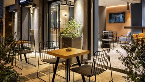 An outdoor seating area with a wooden table and chairs, a potted plant, and a modern interior visible through the glass doors. - Bob W Athens Akadimia (Athens)