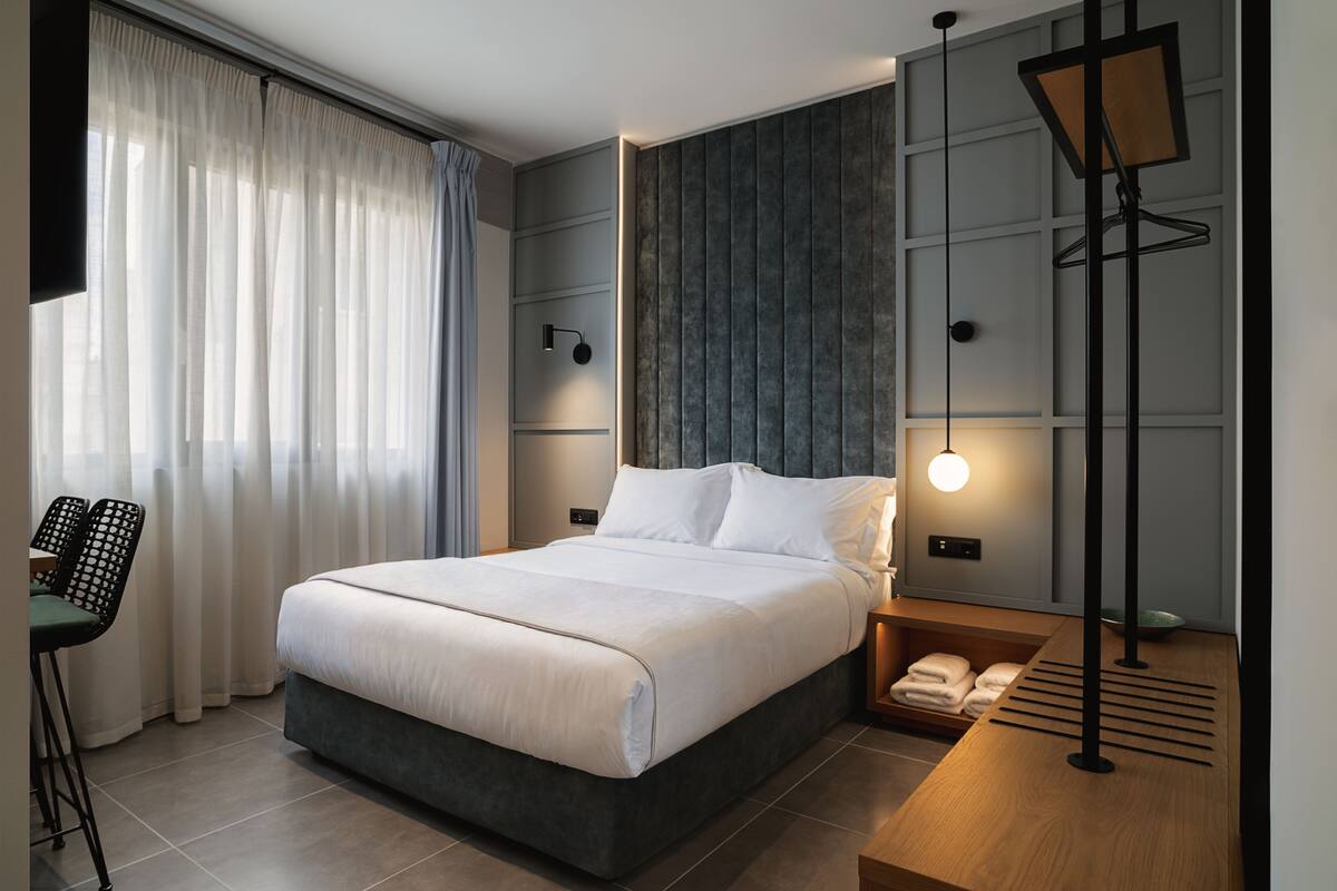 A modern hotel room with a large bed, a desk, a chair, a bedside table, and a wall-mounted lamp.