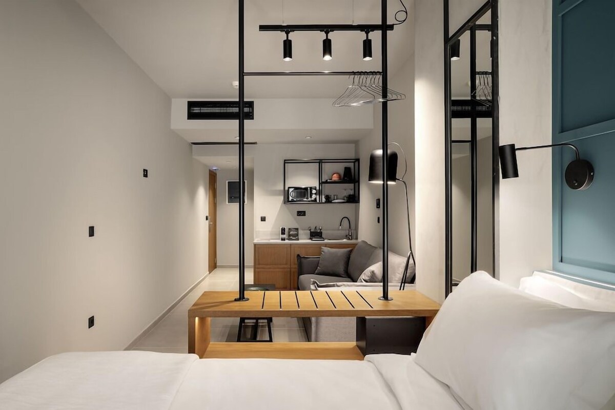 A compact hotel room with a bed, a small table, a kitchenette, and a view of the outside.