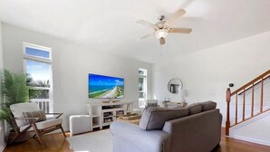 A living room with a grey sofa, a white armchair, a TV, and a ceiling fan. - 1st Tier Beach Home w/ Heated Pool & Gulf Views! (Port St. Joe)