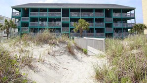 A multi-story building with a flat roof and balconies, surrounded by sand dunes and sparse vegetation.