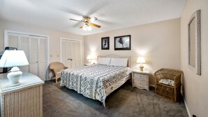 A bedroom with a bed, two bedside lamps, a ceiling fan, a wicker side table, a wicker chair, and two framed pictures on the wall.