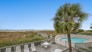 A wooden deck with white lounge chairs and a pool area, surrounded by a white fence and a palm tree.