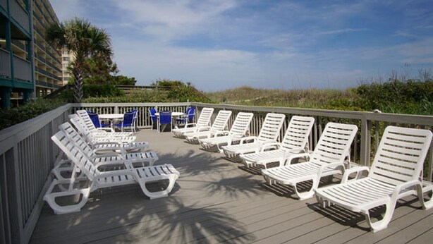 A deck with white lounge chairs and blue chairs, overlooking a beach and palm trees.