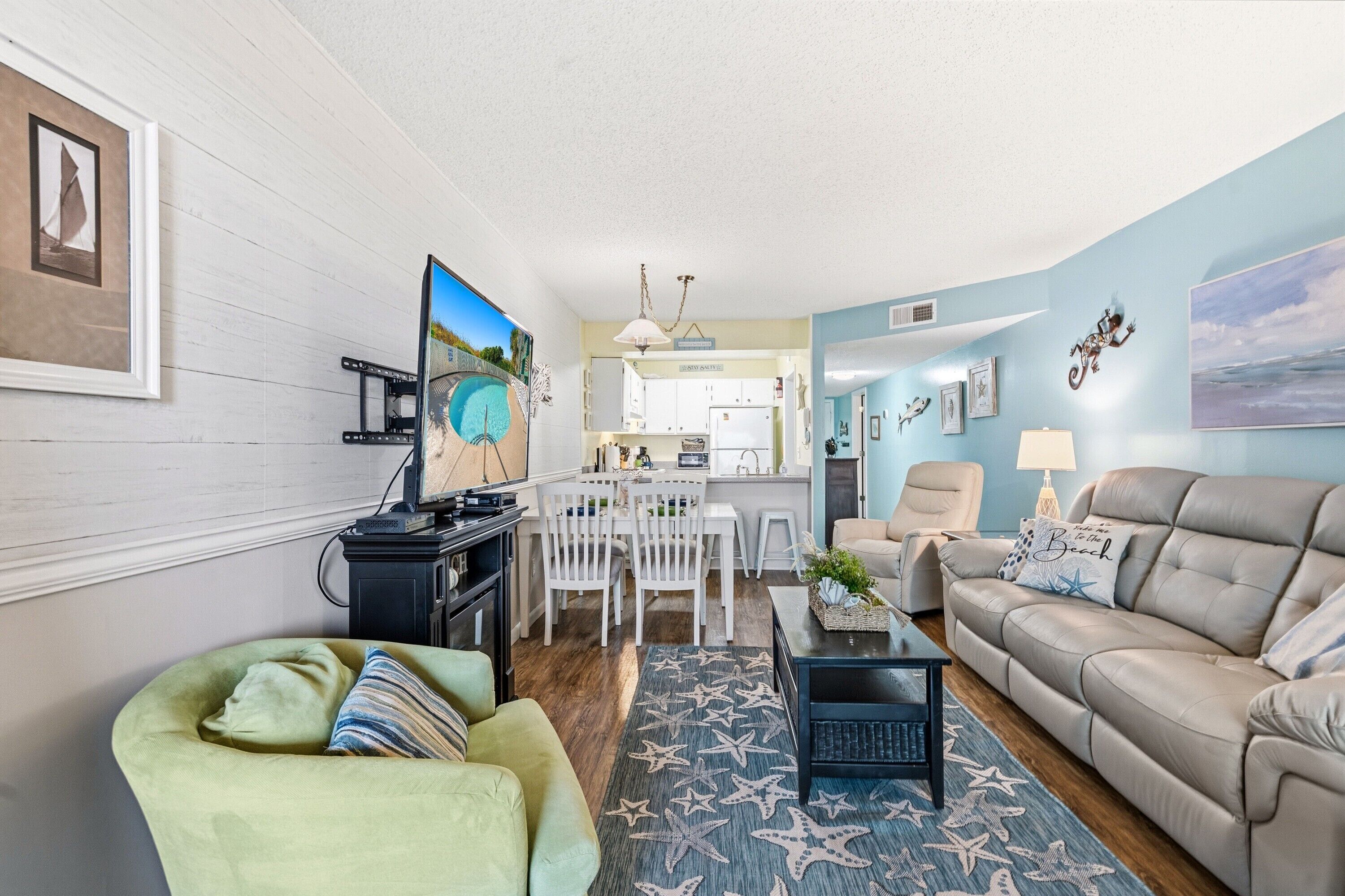 A living room with a sofa, a green armchair, a coffee table, and a TV. There is a dining area with a table and chairs. The room has a light blue wall and a star-patterned rug.