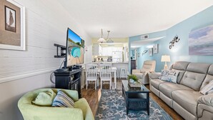 A living room with a sofa, a green armchair, a coffee table, and a TV. There is a dining area with a table and chairs. The room has a light blue wall and a star-patterned rug.