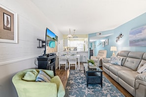 A living room with a sofa, a green armchair, a coffee table, and a TV. There is a dining area with a table and chairs. The room has a light blue wall and a star-patterned rug.