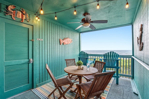 A covered outdoor seating area with a table and chairs, a ceiling fan, string lights, and a view of the ocean.