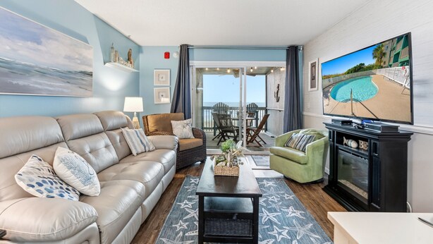 A living room with a leather sofa, a coffee table, and a TV. There is a balcony with a pool view.