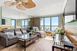 Een ruime woonkamer met een bank, fauteuils, een salontafel en uitzicht op zee door grote ramen.