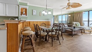 A spacious living room with a dining area, wooden flooring, and a large window with curtains. - Windemere 1006-3BR Oceanfront Ocean Drive (North Myrtle Beach)