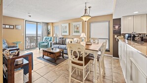 A spacious living room with a dining area, a kitchen, and a sliding glass door leading to a balcony. - Waterpointe II 806-3BR/2BA Oceanfront Condo in Windy Hill (North Myrtle Beach)