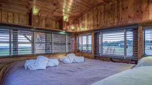 A wooden cabin room with a large bed, two windows with blinds, and a view of the outdoors.