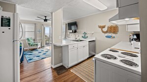 A modern kitchen with white cabinets, a stove, and a sink. There is a ceiling fan and a television mounted on the wall. - Windsong 106-1BR/1BA Oceanfront condo in Windy Hill (North Myrtle Beach)