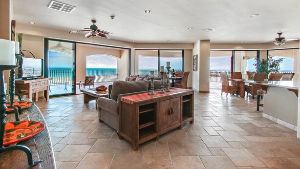 A spacious living area with a large window overlooking the ocean, a dining area with wicker furniture, and a ceiling fan. - Luxury Three Bedroom Ocean Front Condominium (San Felípe)