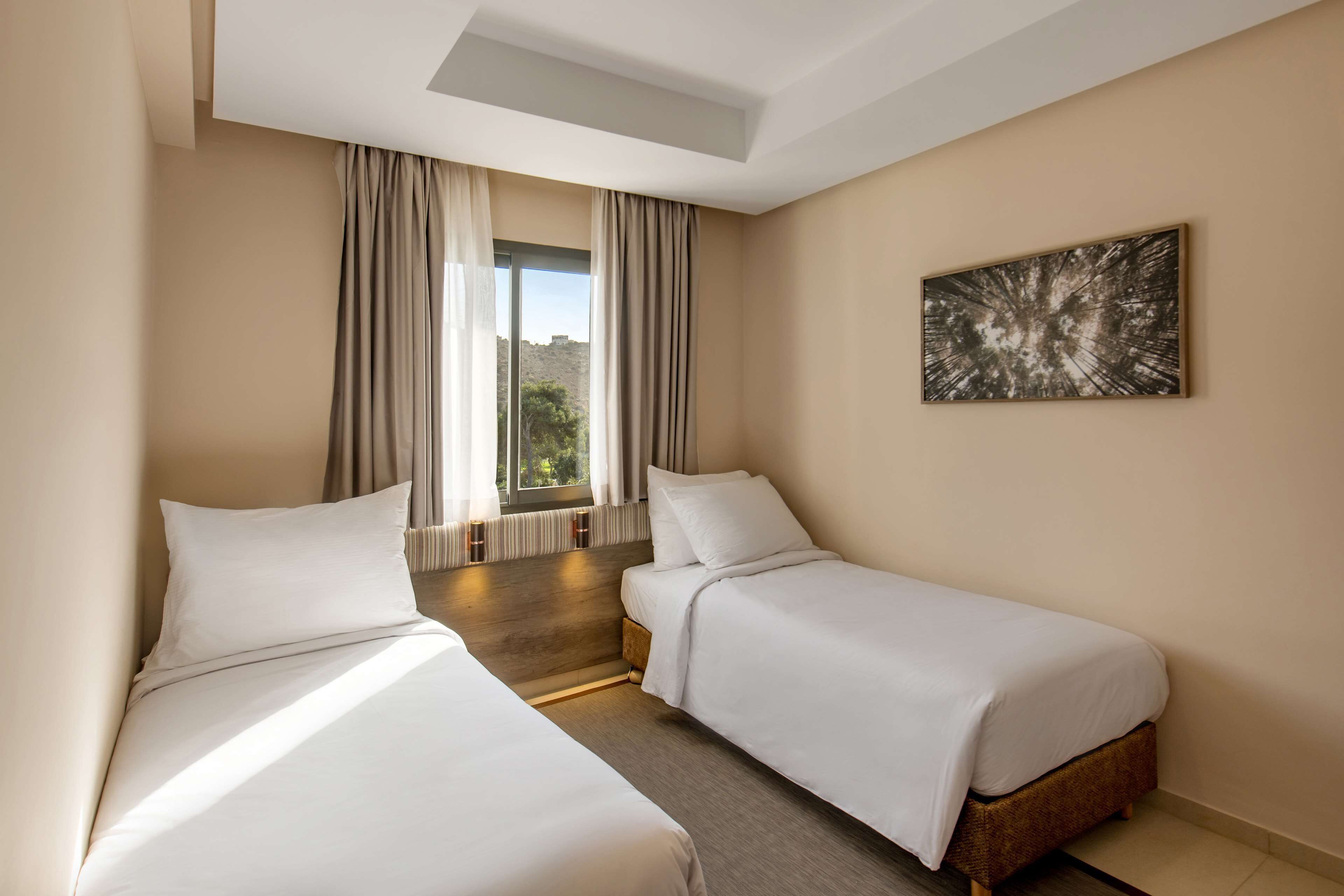 Une chambre d’hôtel avec deux lits simples, une grande fenêtre avec des rideaux, un luminaire au plafond et une œuvre d’art abstraite accrochée au mur.