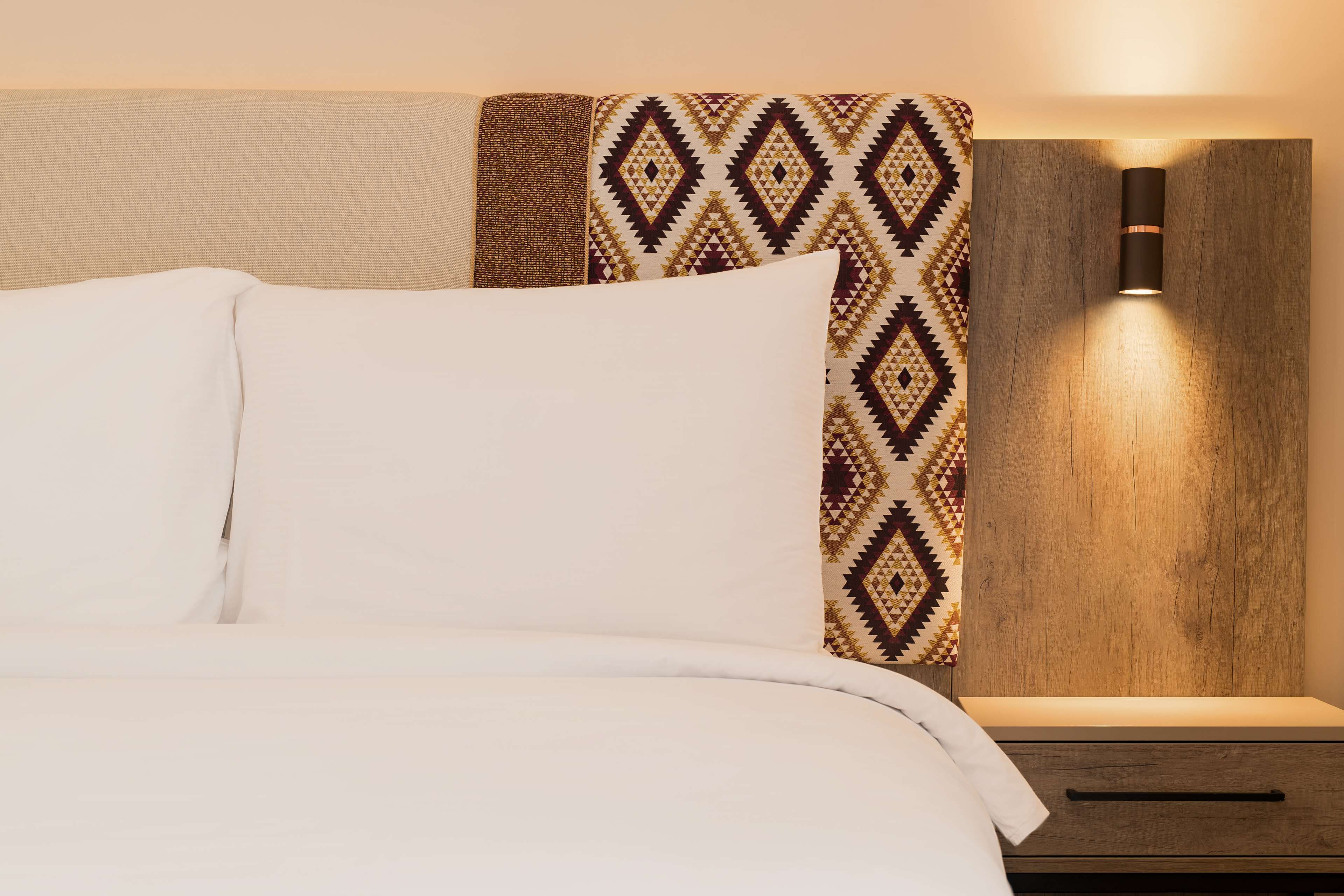 A neatly made bed with white linens, a patterned headboard, and a wall-mounted light fixture.